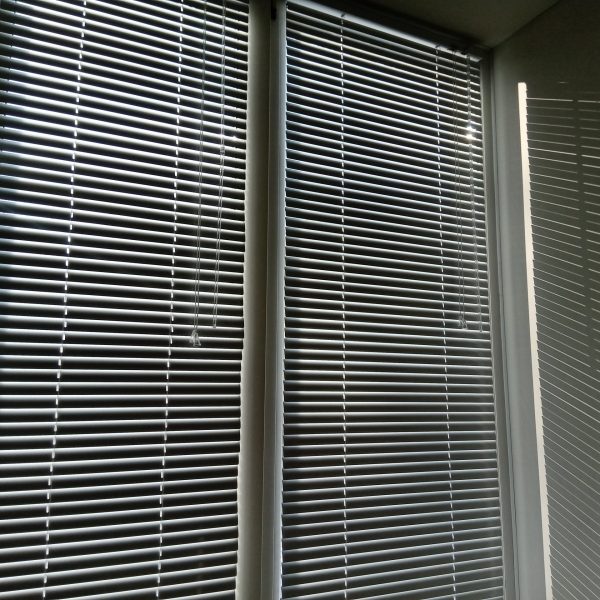 White horizontal blinds on the window block out the bright sunlight. shadow from the blinds